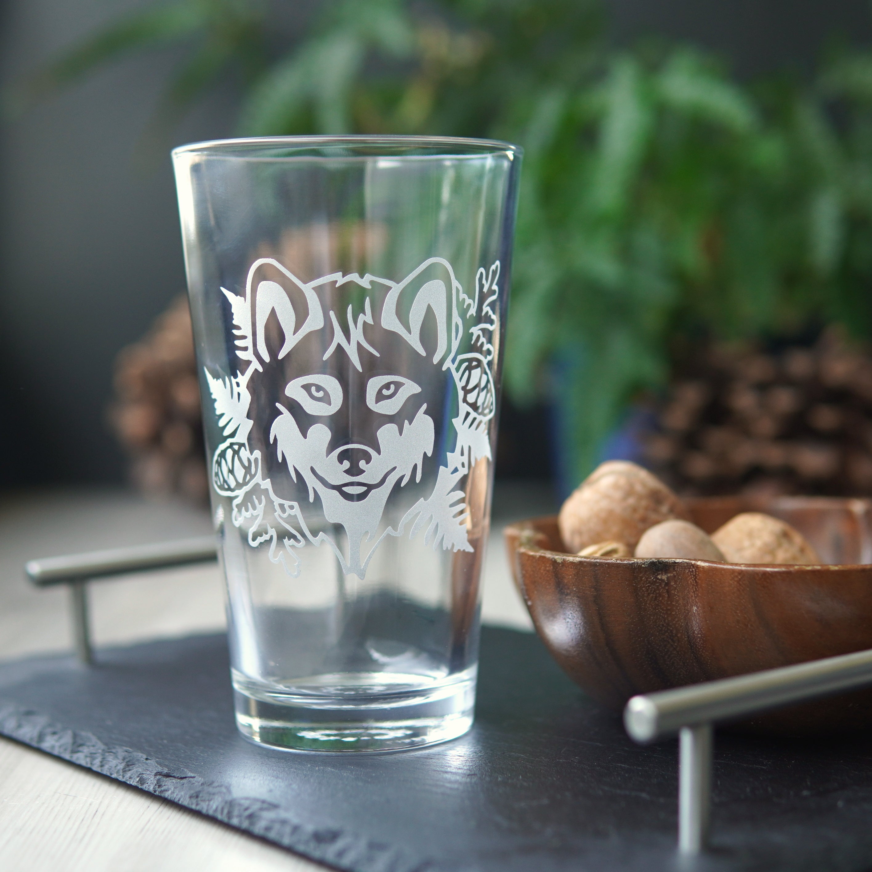 Pint glass engraved with a wolf's face, ferns, cedar branches, and pine cones. The glass is sitting on a black slate tray, next to a wooden bowl of nuts, with ferns and pine cones in the background.