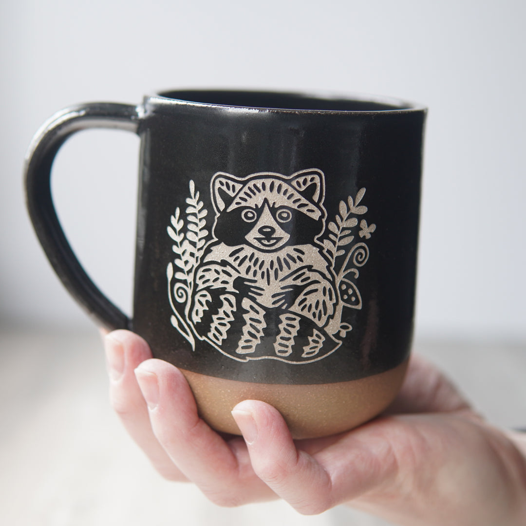 black mug, engraved with a smiling raccoon, held by a hand