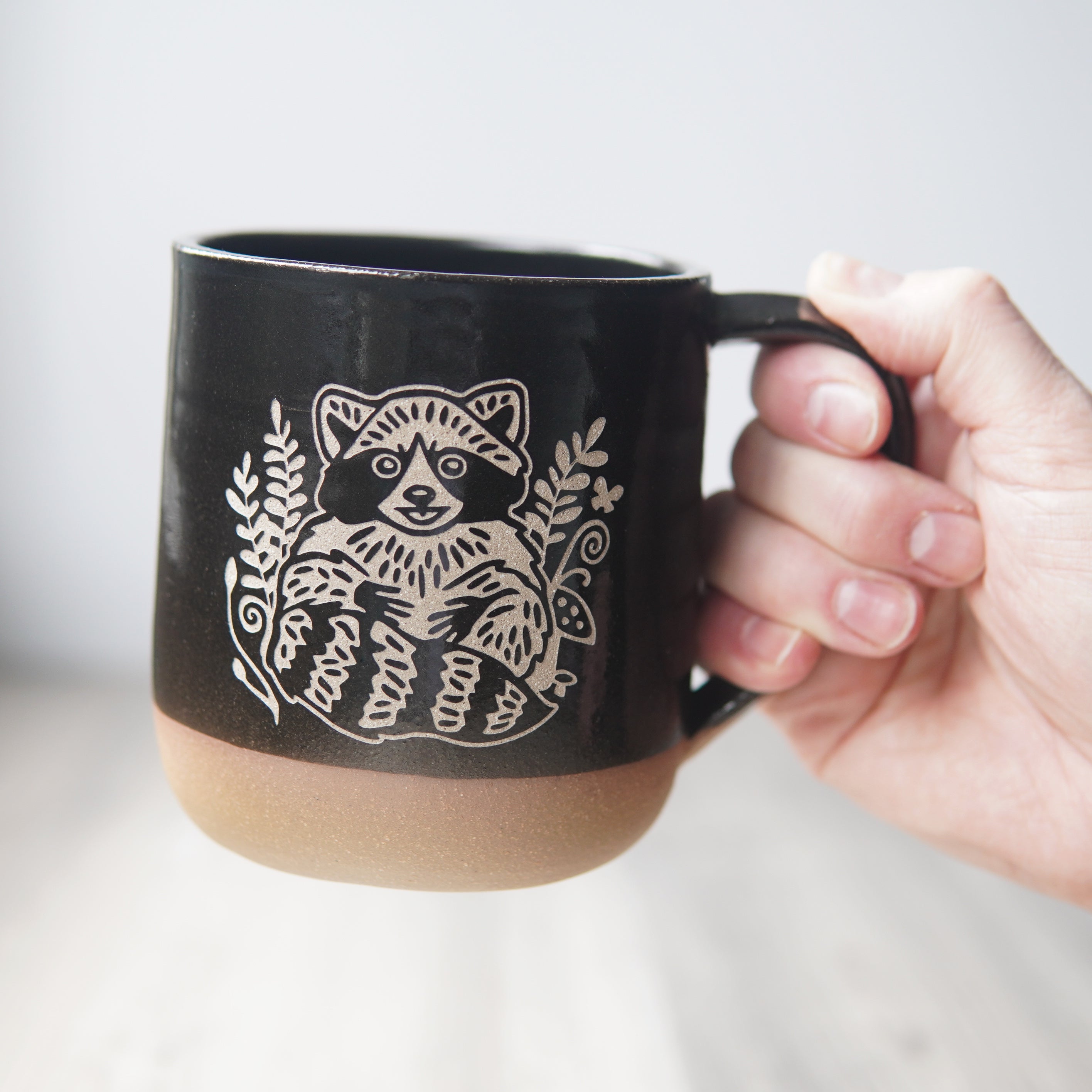 black mug, engraved with a smiling raccoon, held by a hand