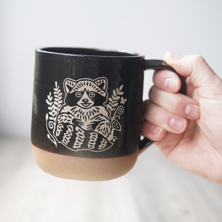 black mug, engraved with a smiling raccoon, held by a hand