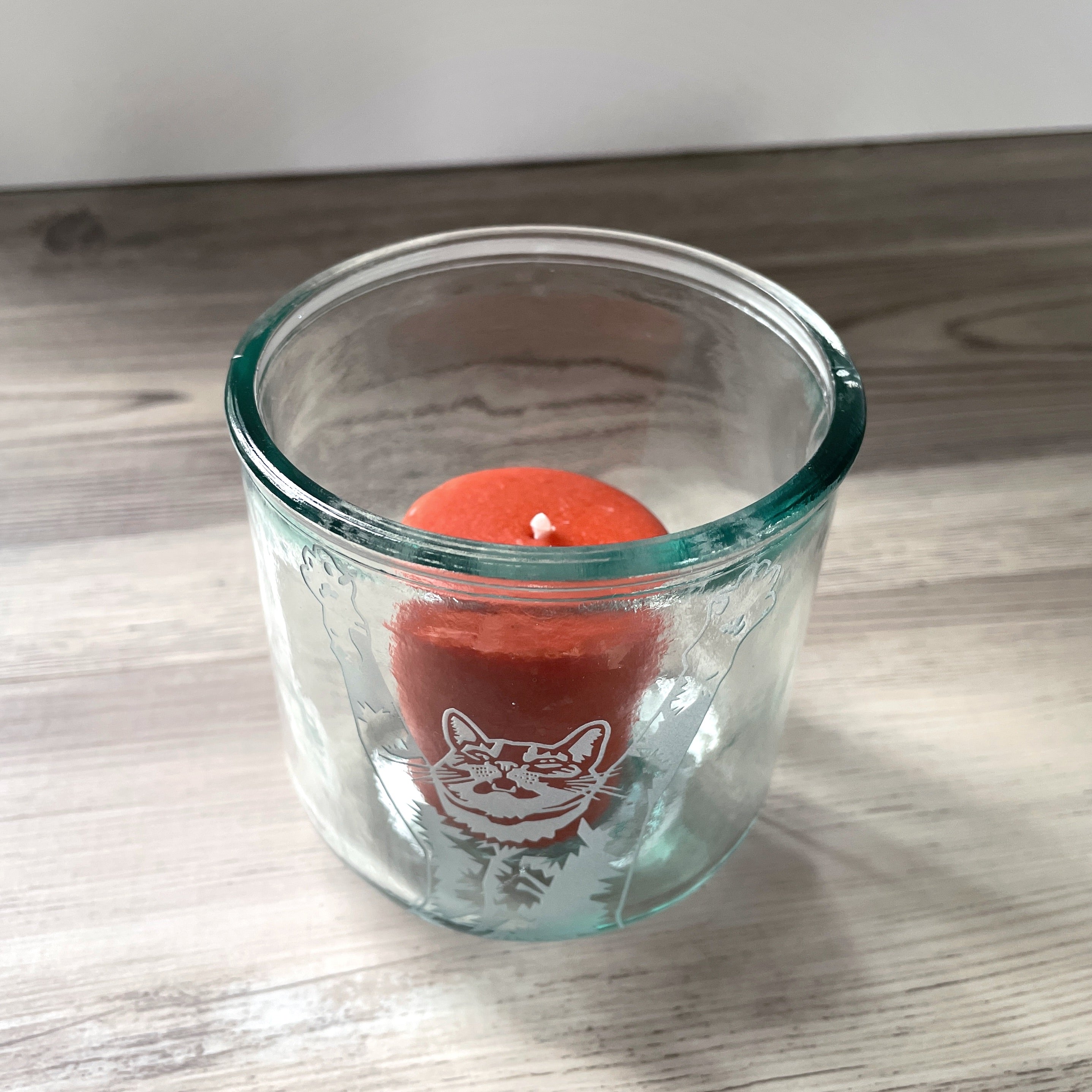top half of a tabby cat with its arms stretched up and mouth slightly open engraved onto a short glass holding a votive candle