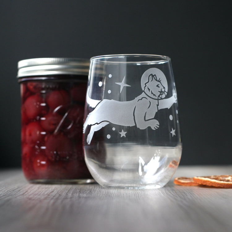 an astronaut cat reaching for the stars, engraved onto a stemless wine glass. There are orange slices and a jar of brandied cherries in the background.