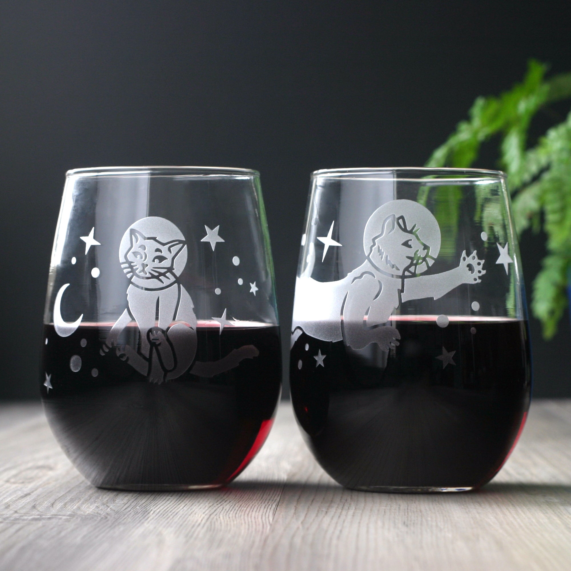 space cats engraved onto a pair of stemless wine glasses. There is red wine in both glasses.