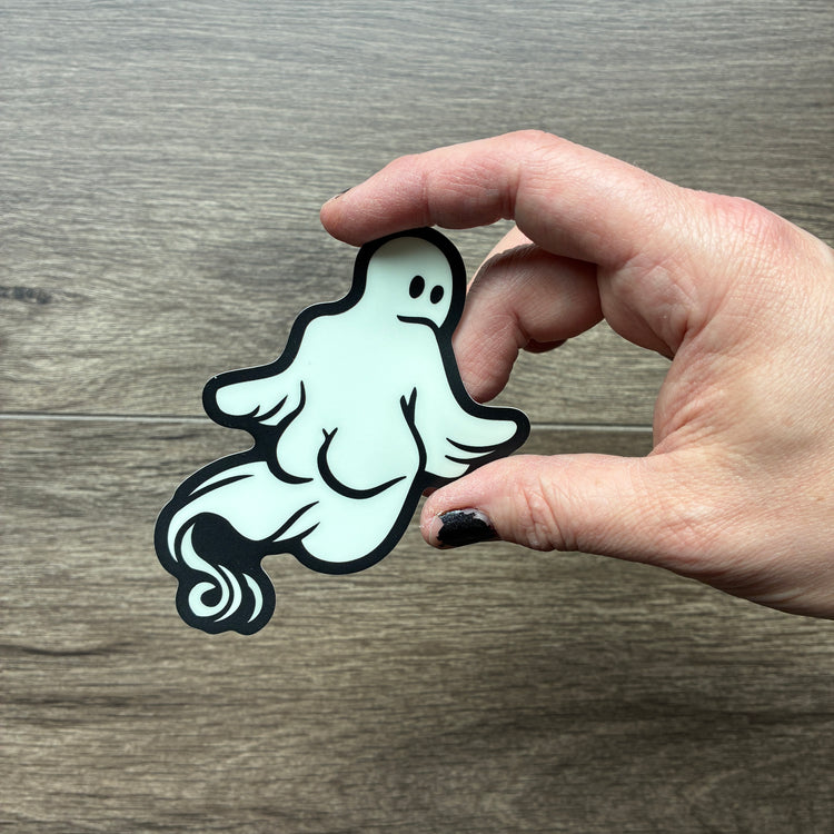 A hand holding a die-cut sticker of a ghost looking over its shoulder at you with a black outline and white body, set against a gray wood-grain background. The ghost has a very curvy backside, with well-defined butt-cheeks.