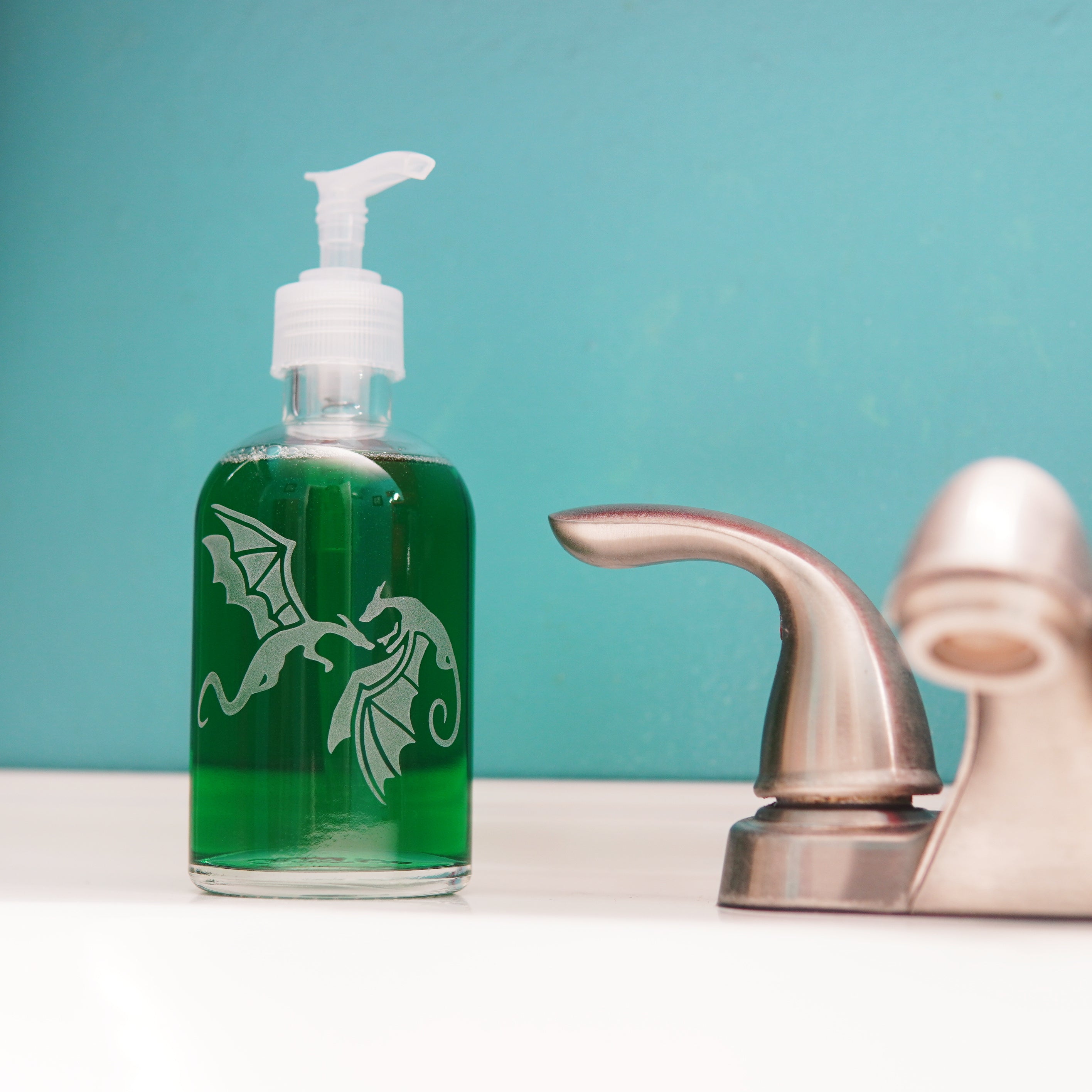 glass pump bottle soap dispenser full of green soap next to a bathroom faucet, with a pair of flying dragons engraved on it