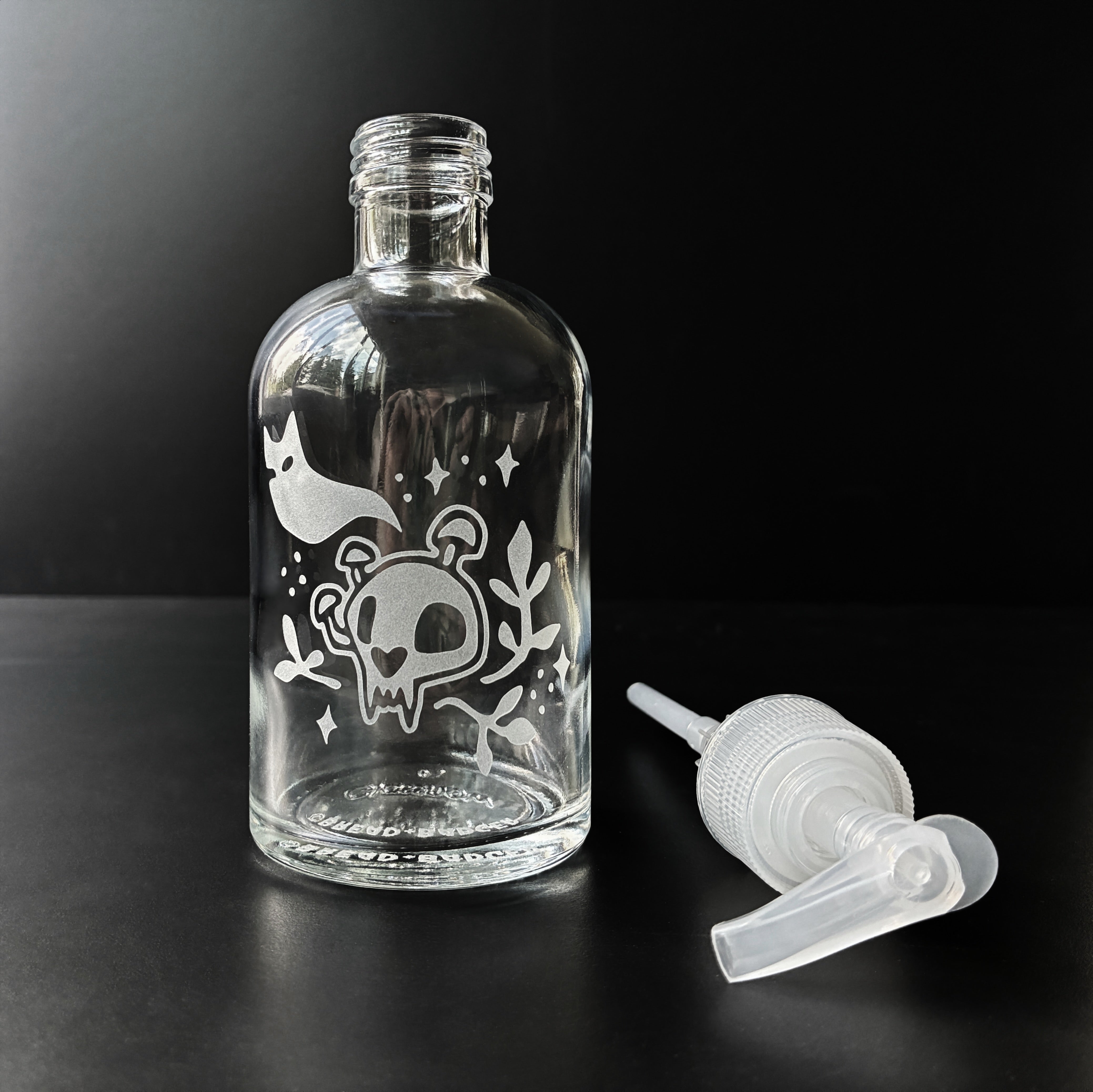 Clear glass bottle with decorative etching of a ghost and cat skull on a black background