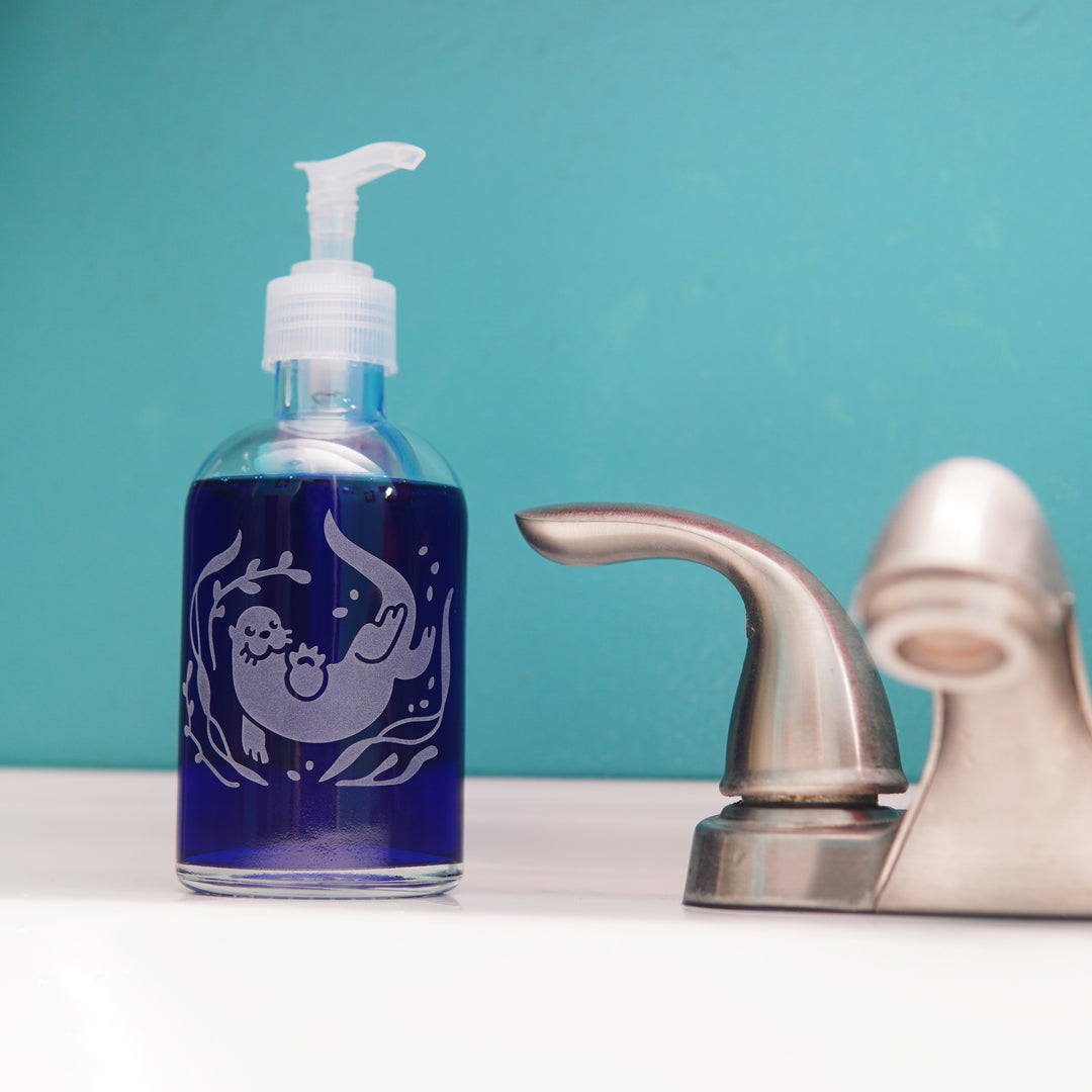 glass pump bottle soap dispenser full of dark blue soap next to a bathroom faucet, with an otter engraved on it