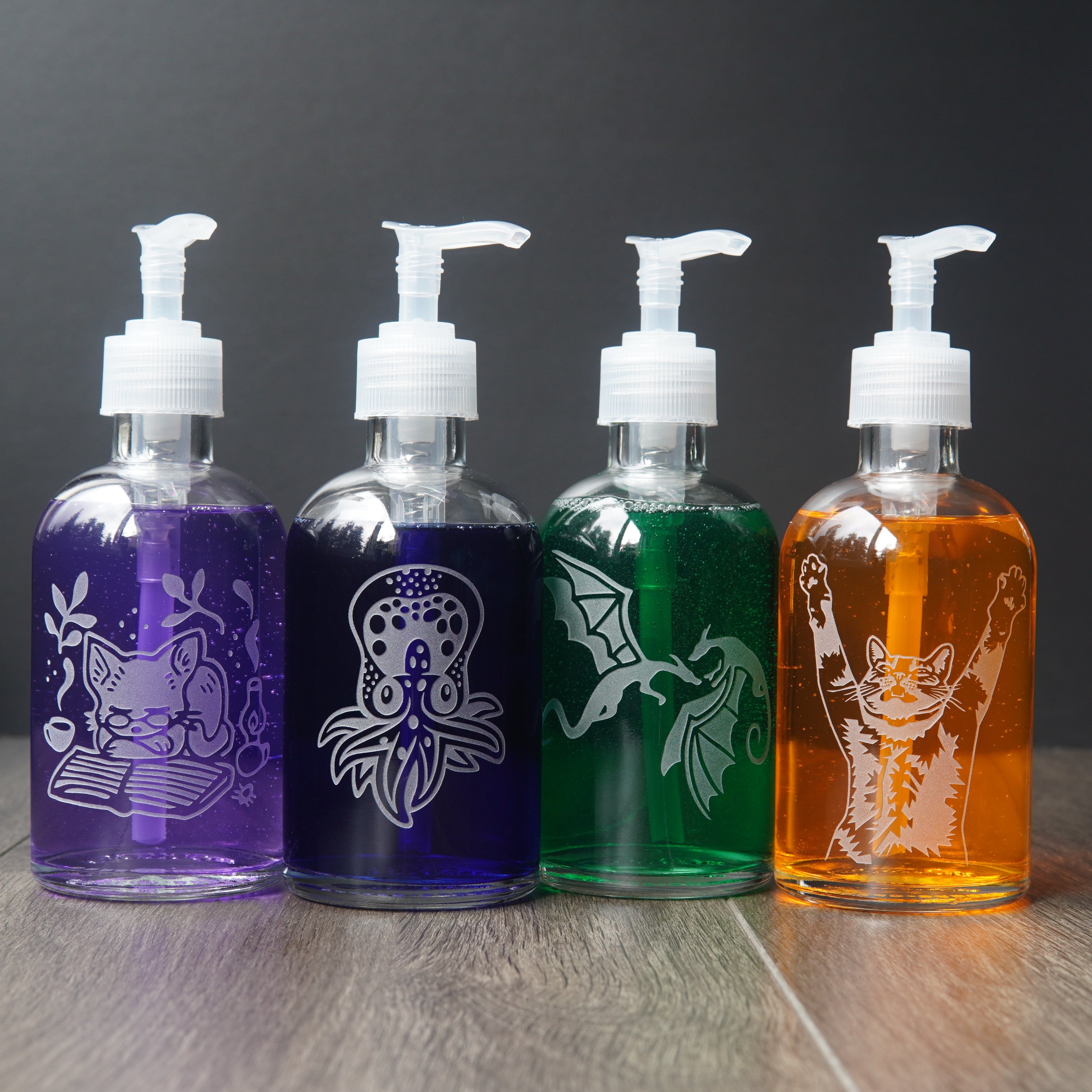 4 glass soap dispensers with different colored soap in them, and different animals engraved on them. From left to right: purple cat with book, dark blue octopus, green flying dragons, and orange cat with arms up