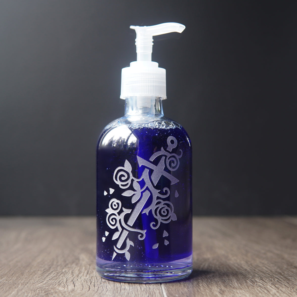 glass soap dispenser engraved with a sword wrapped with a rose vine. the pump bottle is filled with dark blue liquid soap
