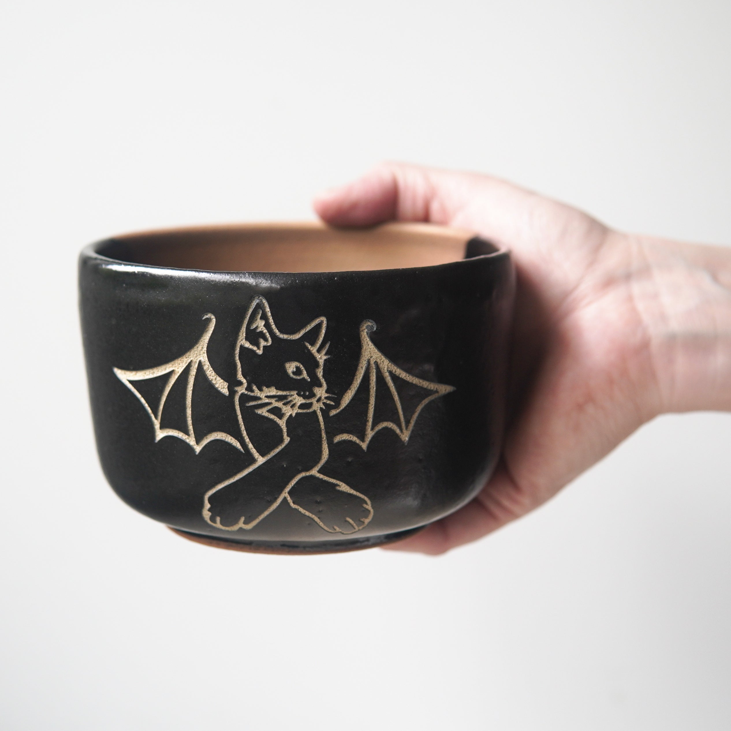 Cat Bat engraved onto a black pottery bowl, held by a hand