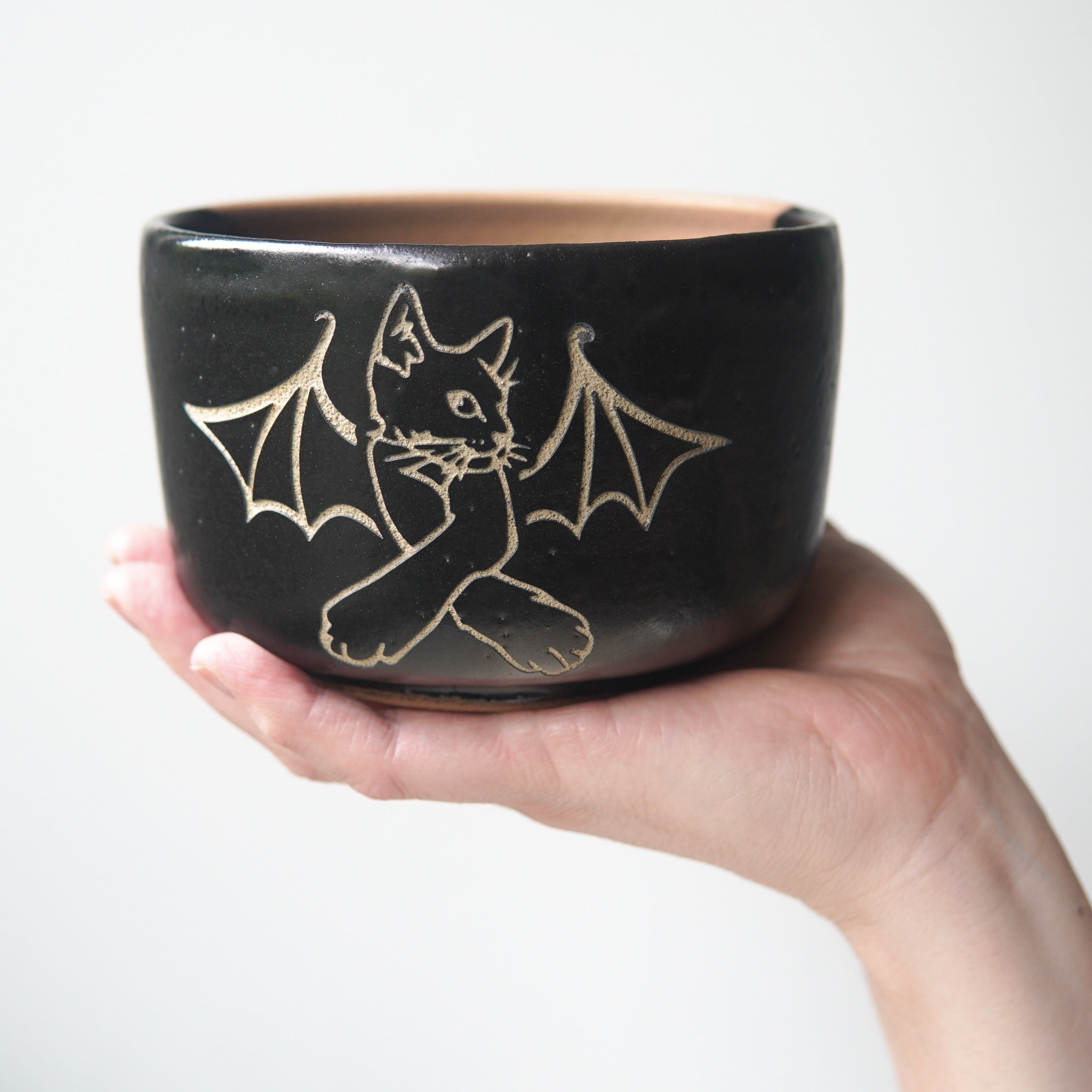 Cat Bat engraved onto a black pottery bowl, held by a hand