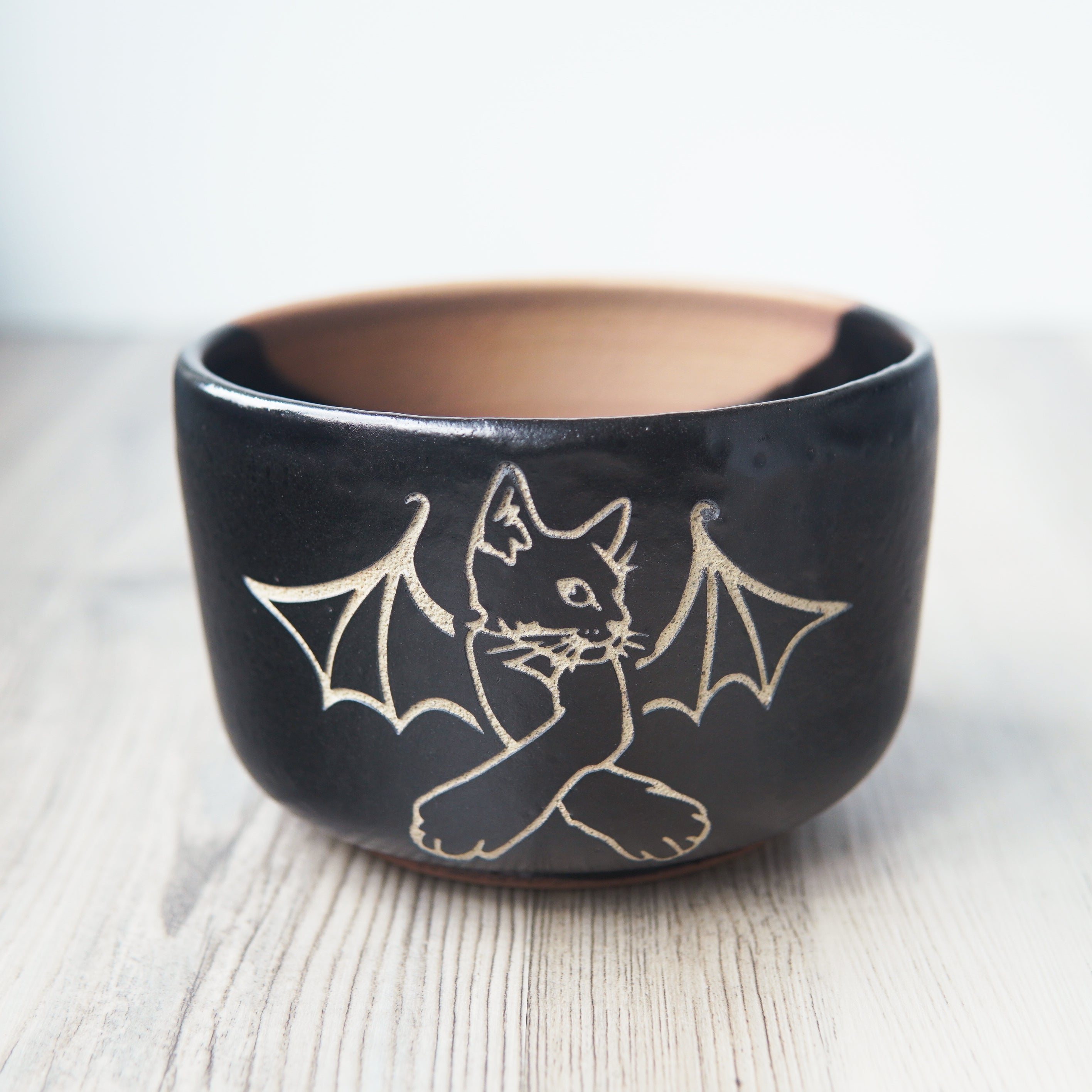 Cat Bat engraved onto a black pottery bowl. There's an exposed area on the far side of the bowl which isn't glazed.
