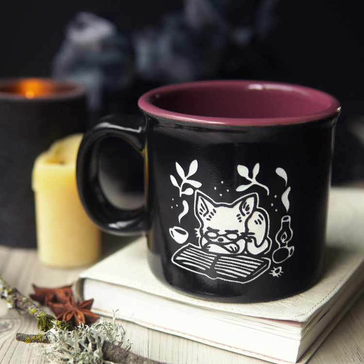 Black mug with a white cat illustration on a book on a wooden surface with candles in the background