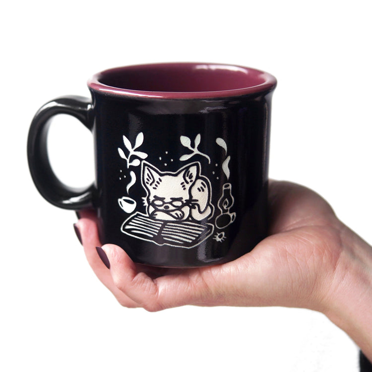 Black mug with white cat illustration held by a hand on a white background