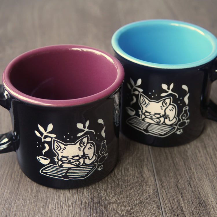 Two black mugs with cat illustrations on a wooden surface
