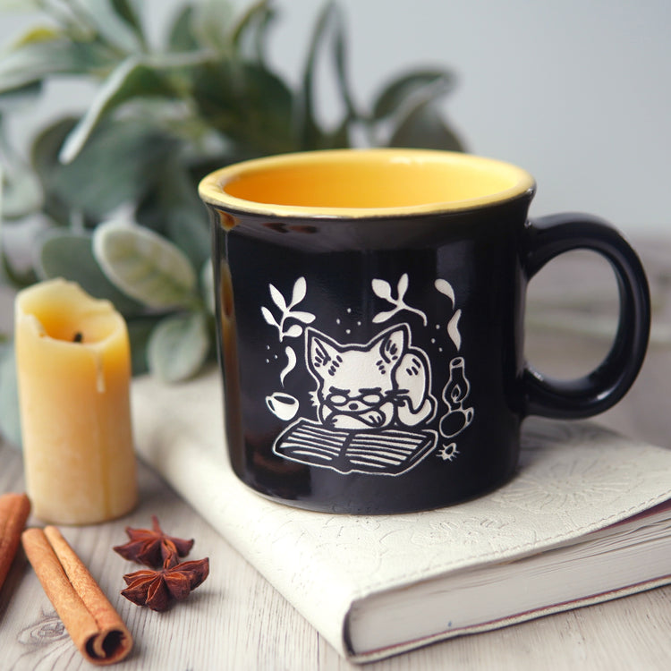 Black mug with a white cat illustration on a book on a wooden surface with candles in the background