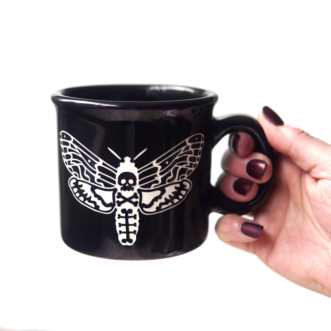 Black mug with white deaths head hawkmoth design held by a hand on a white background