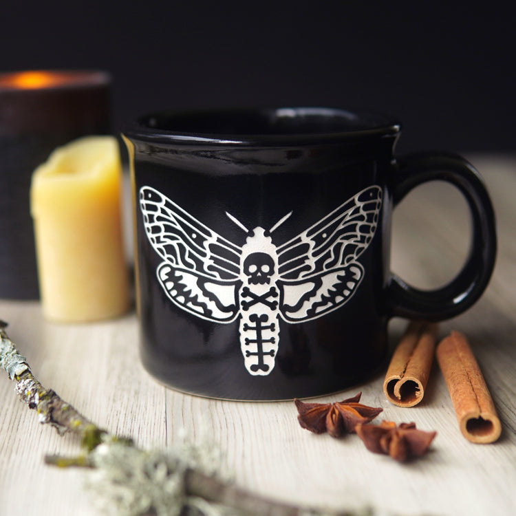 Black mug with white deaths head hawkmoth design on a wooden surface with candles, a mossy stick, cinnamon sticks, and star anise in the background.