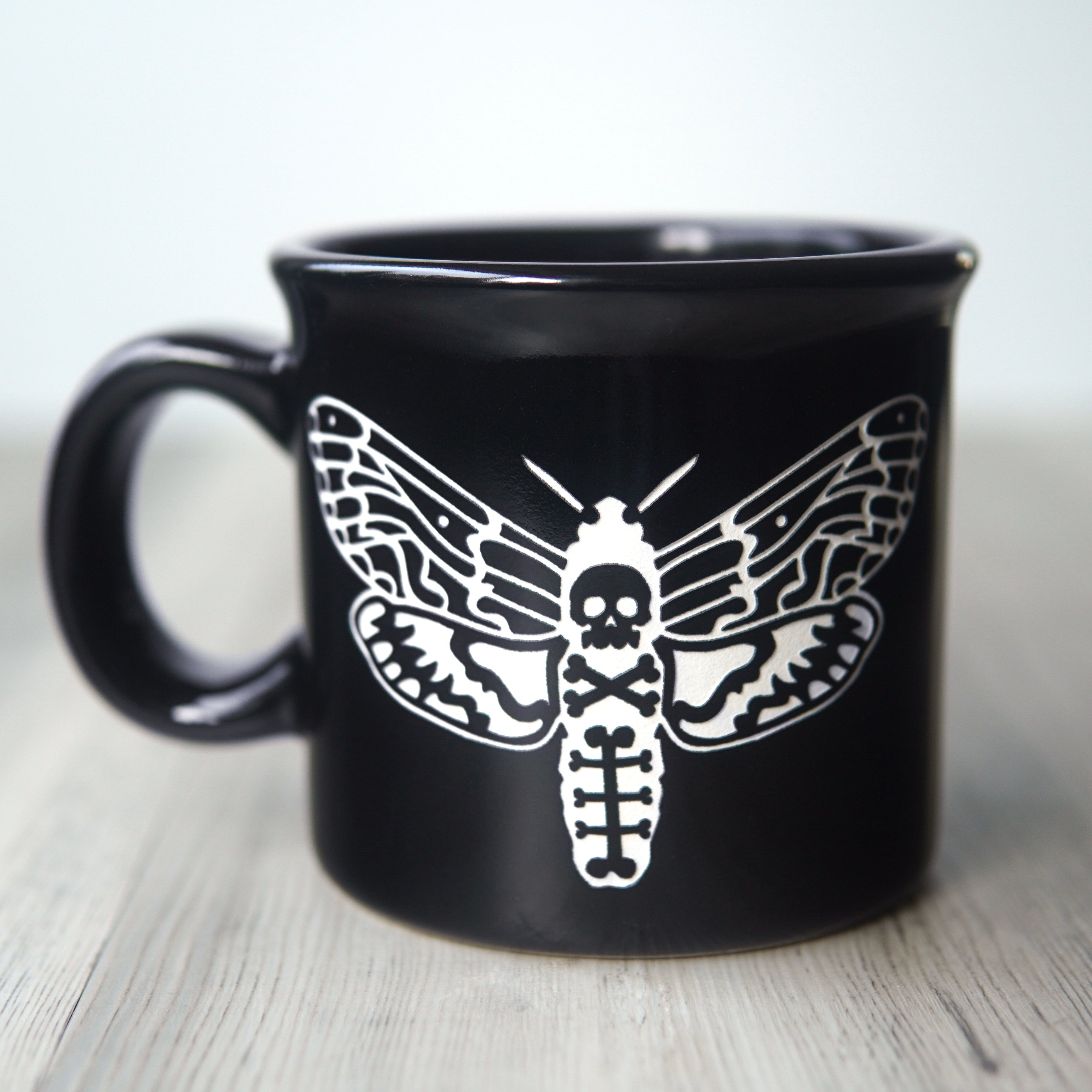 Black mug with white deaths head hawkmoth design on a wooden surface