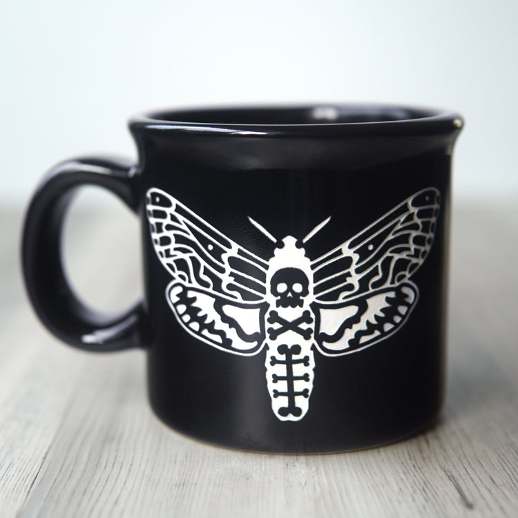 Black mug with white deaths head hawkmoth design on a wooden surface