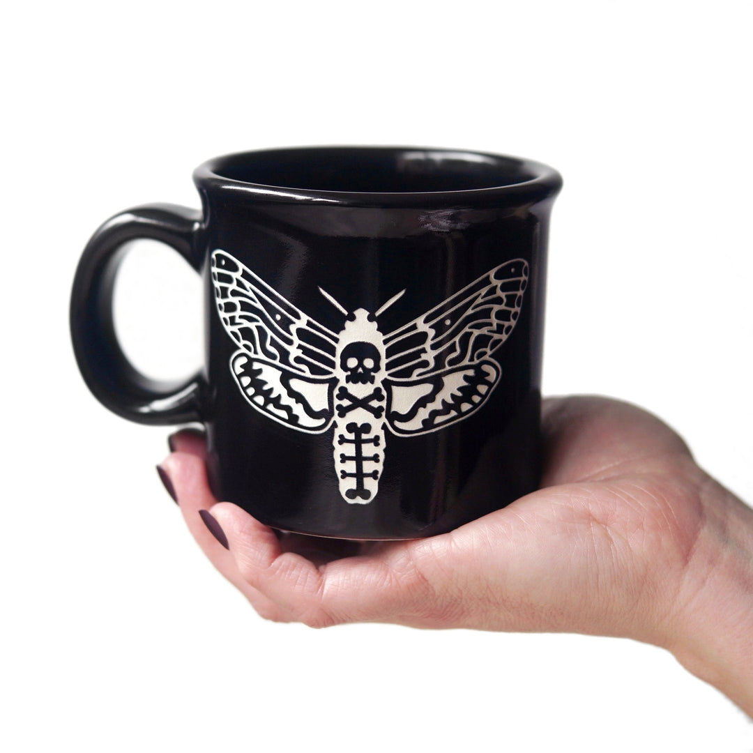 Black mug with white deaths head hawkmoth design held by a hand on a white background