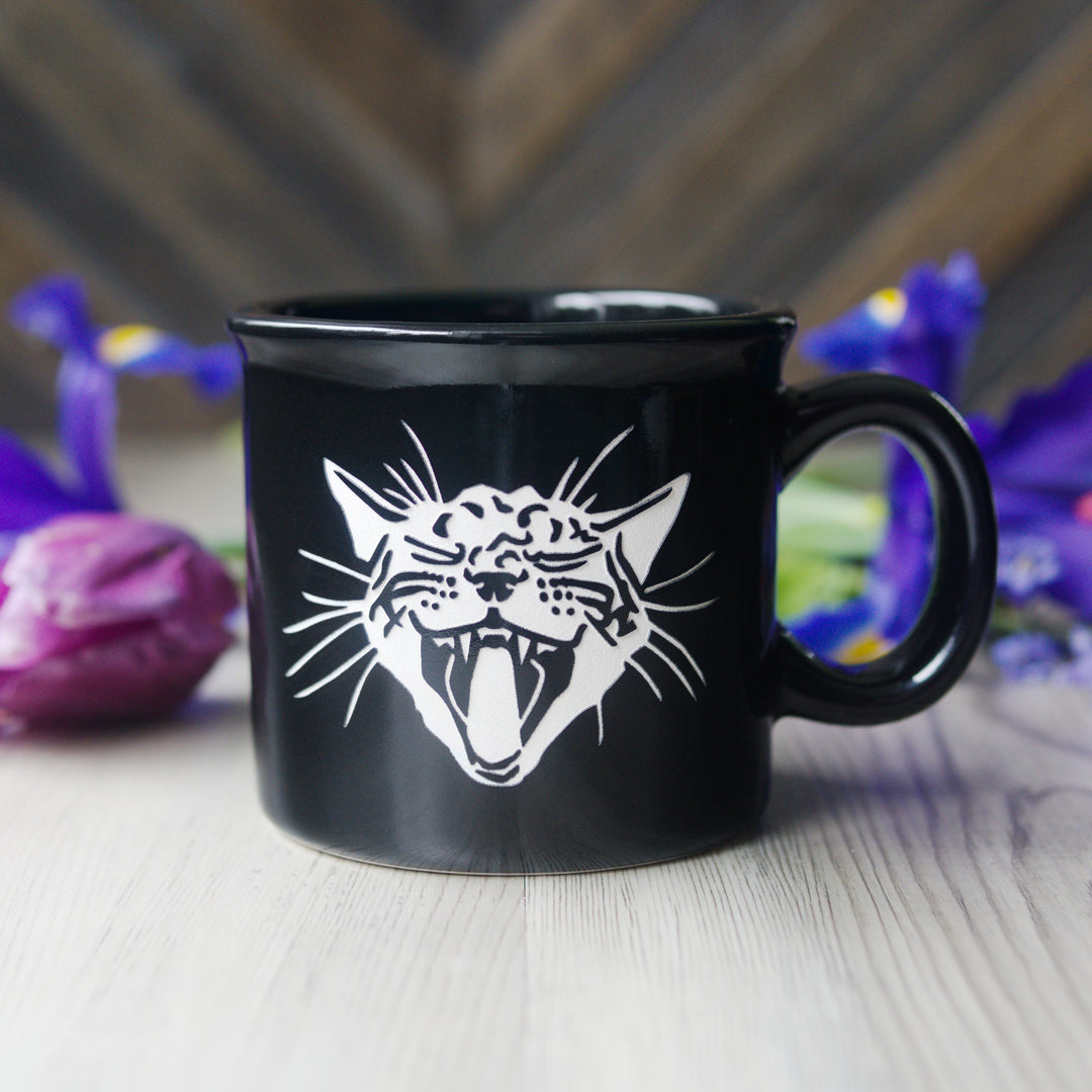 Black mug with a white cat illustration on a wooden surface with blurred flowers in the background