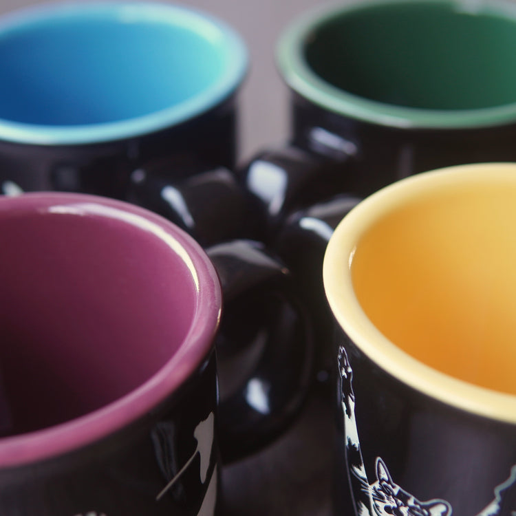 Four black mugs with different colored interiors on a dark surface. Colors are light blue, dark green, burgundy, and yellow.