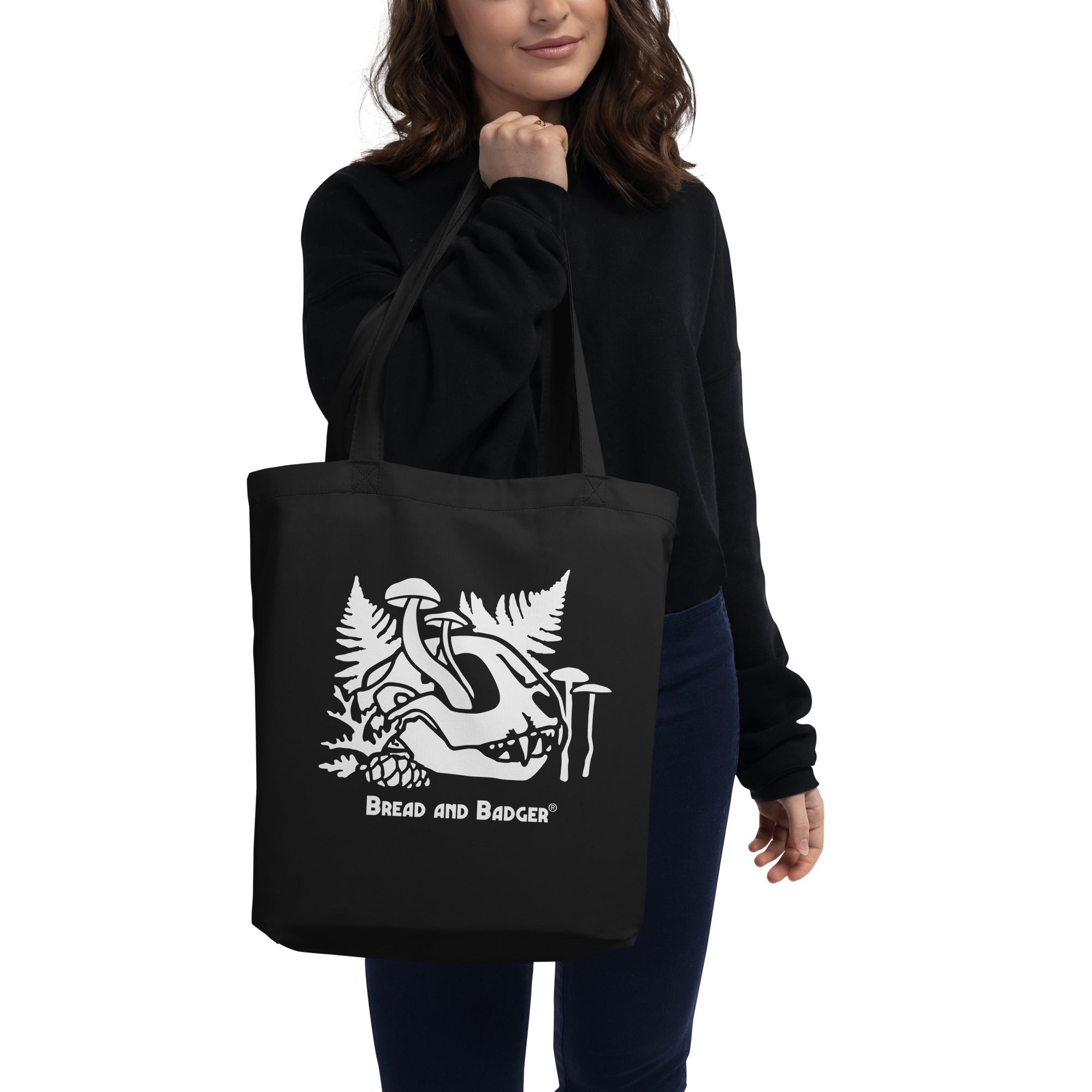 Person holding a black tote bag with a white cat skull and mushrooms design and text on a white background