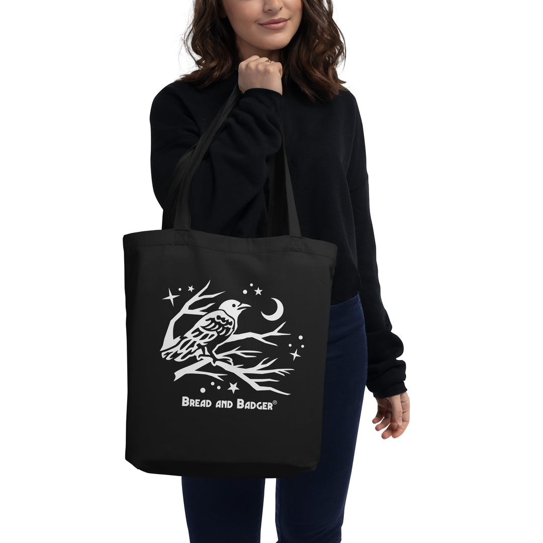 Person holding a black tote bag with a white crow design and 'Bread and Badger' logo.