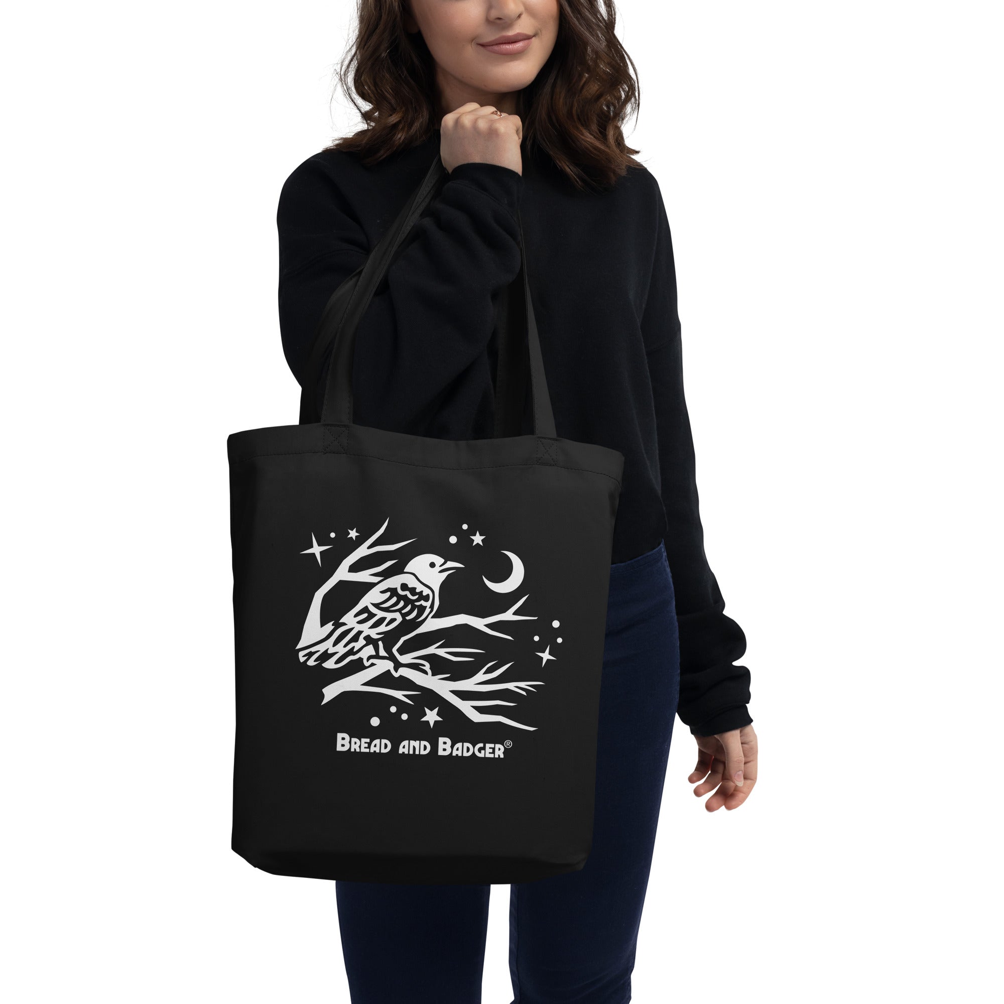 Person holding a black tote bag with a white crow design and 'Bread and Badger' logo.