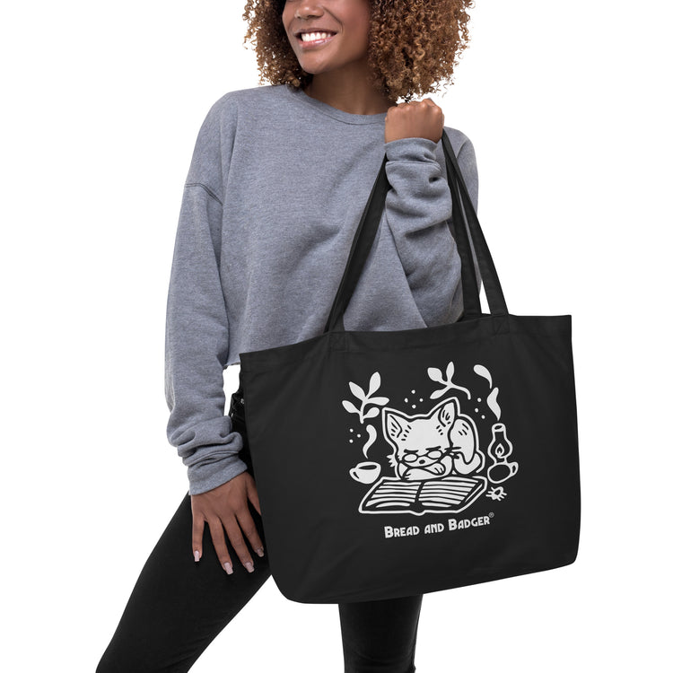 Woman holding a black tote bag with a white cat illustration and 'Bread and Badger' branding on a white background. The cat is sleeping in front of an open book, next to a steaming mug and lantern.