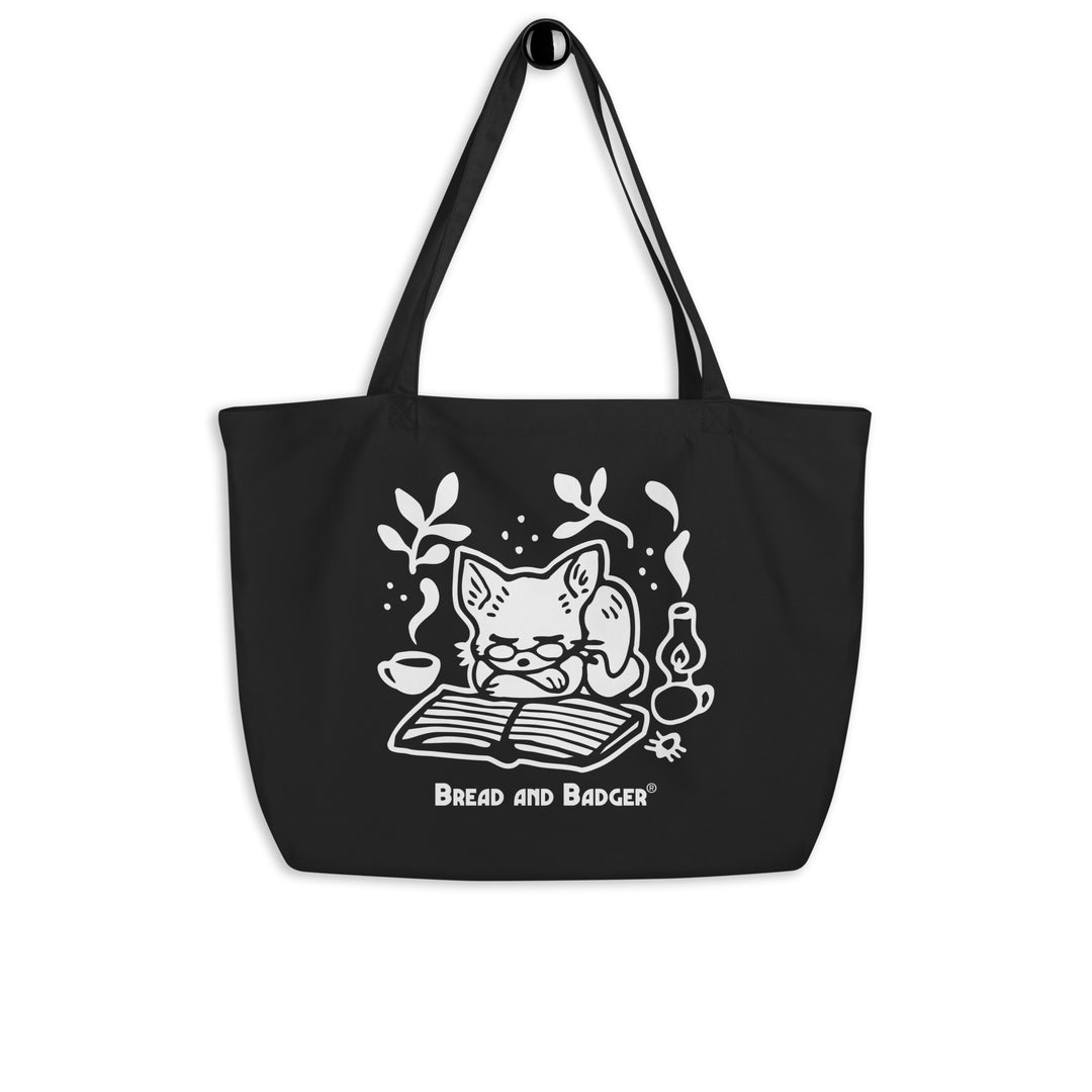 Black tote bag with a white cat illustration and 'Bread and Badger' branding on a white background. The cat is sleeping in front of an open book, next to a steaming mug and lantern.