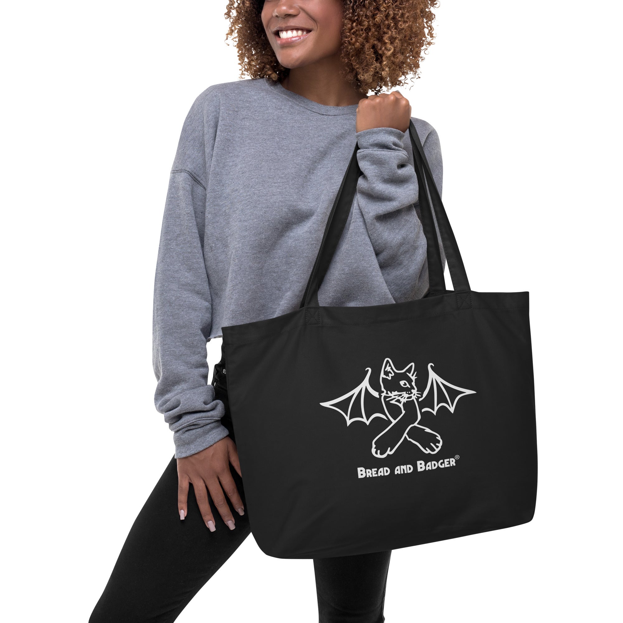 Person holding a black tote bag with a white bat cat design and 'Bread and Badger' text on a white background