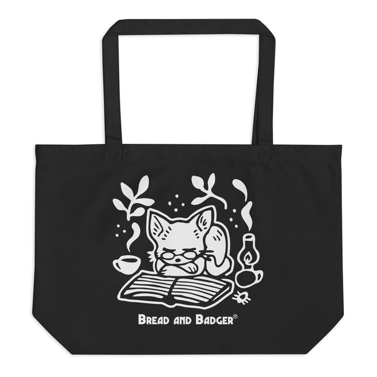 Black tote bag with a white cat illustration and 'Bread and Badger' branding on a white background. The cat is sleeping in front of an open book, next to a steaming mug and lantern.