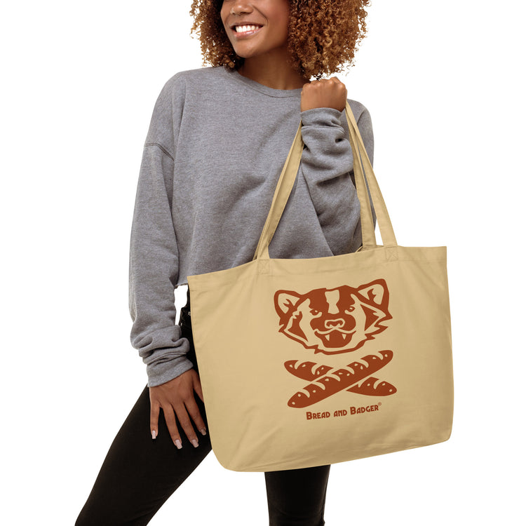 Woman holding a beige tote bag with a red logo and "Bread and Badger" text on a white background. The logo is a badger's face with mouth open above two crossed baguettes.
