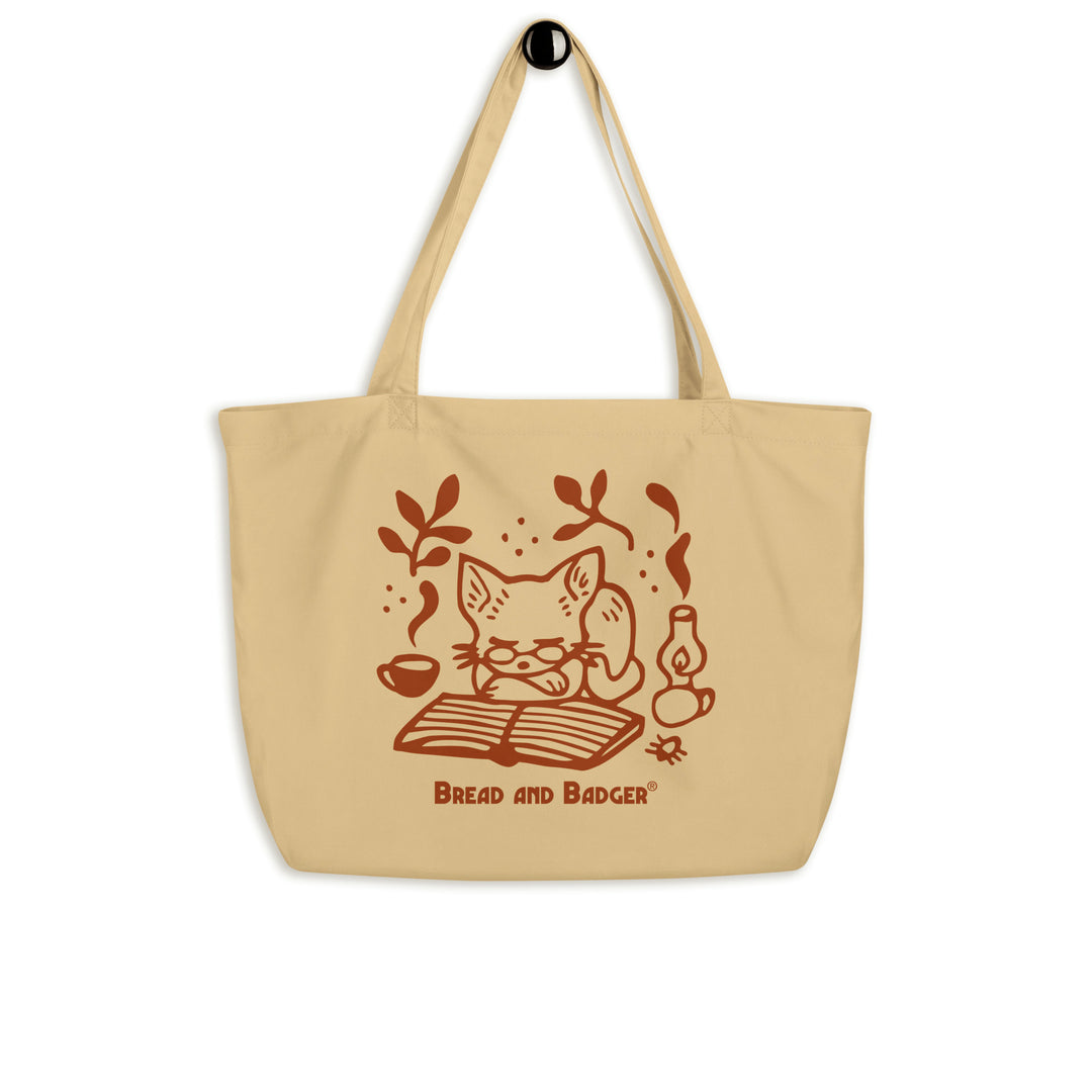 Beige tote bag with a red cat illustration and 'Bread and Badger' branding on a white background. The cat is sleeping in front of an open book, next to a steaming mug and lantern.