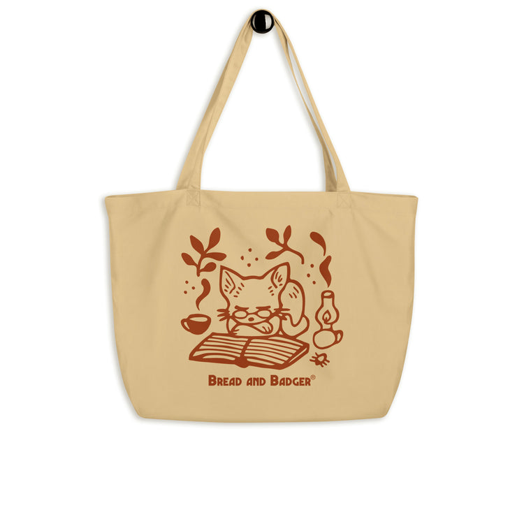 Beige tote bag with a red cat illustration and 'Bread and Badger' branding on a white background. The cat is sleeping in front of an open book, next to a steaming mug and lantern.