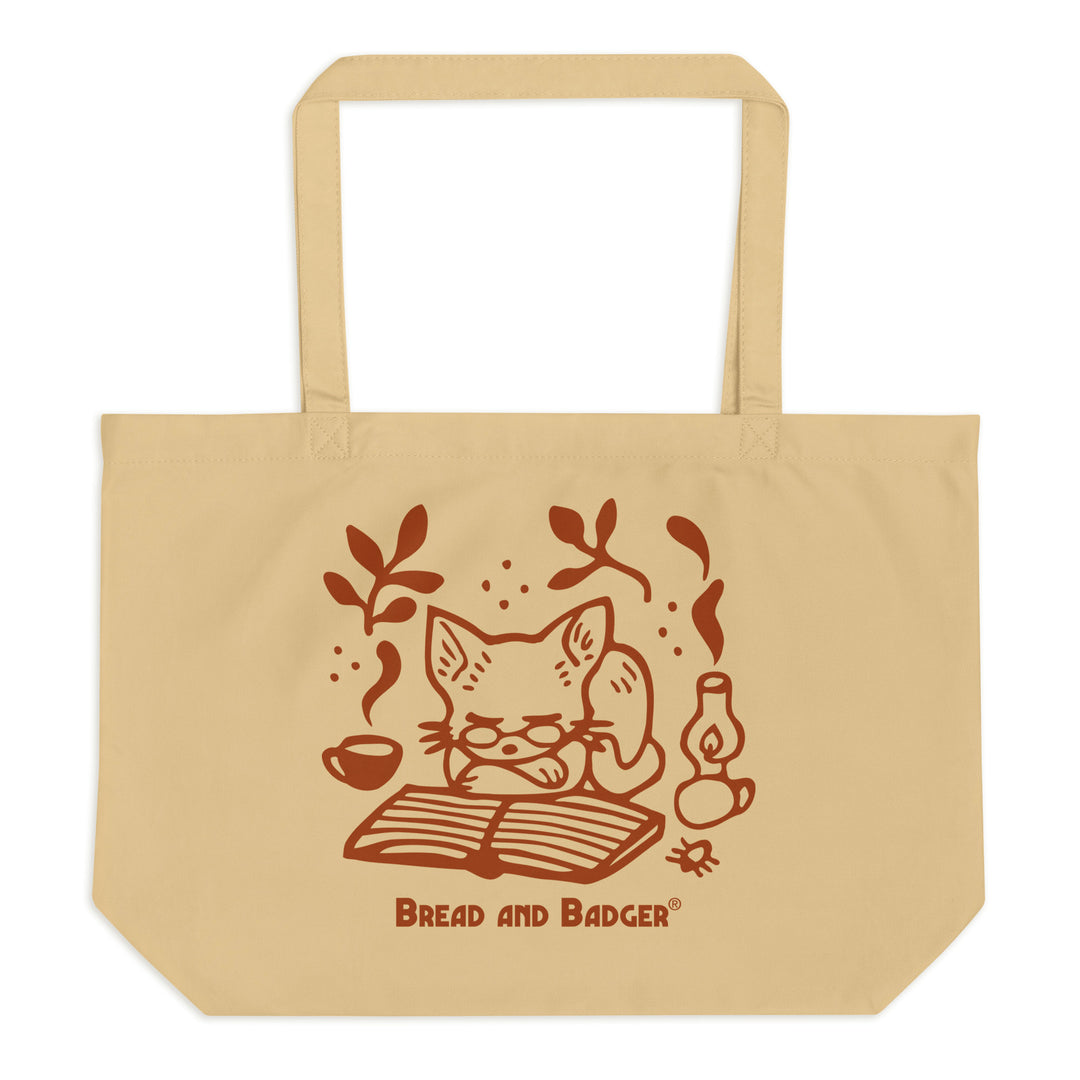 Beige tote bag with a red cat illustration and 'Bread and Badger' branding on a white background. The cat is sleeping in front of an open book, next to a steaming mug and lantern.