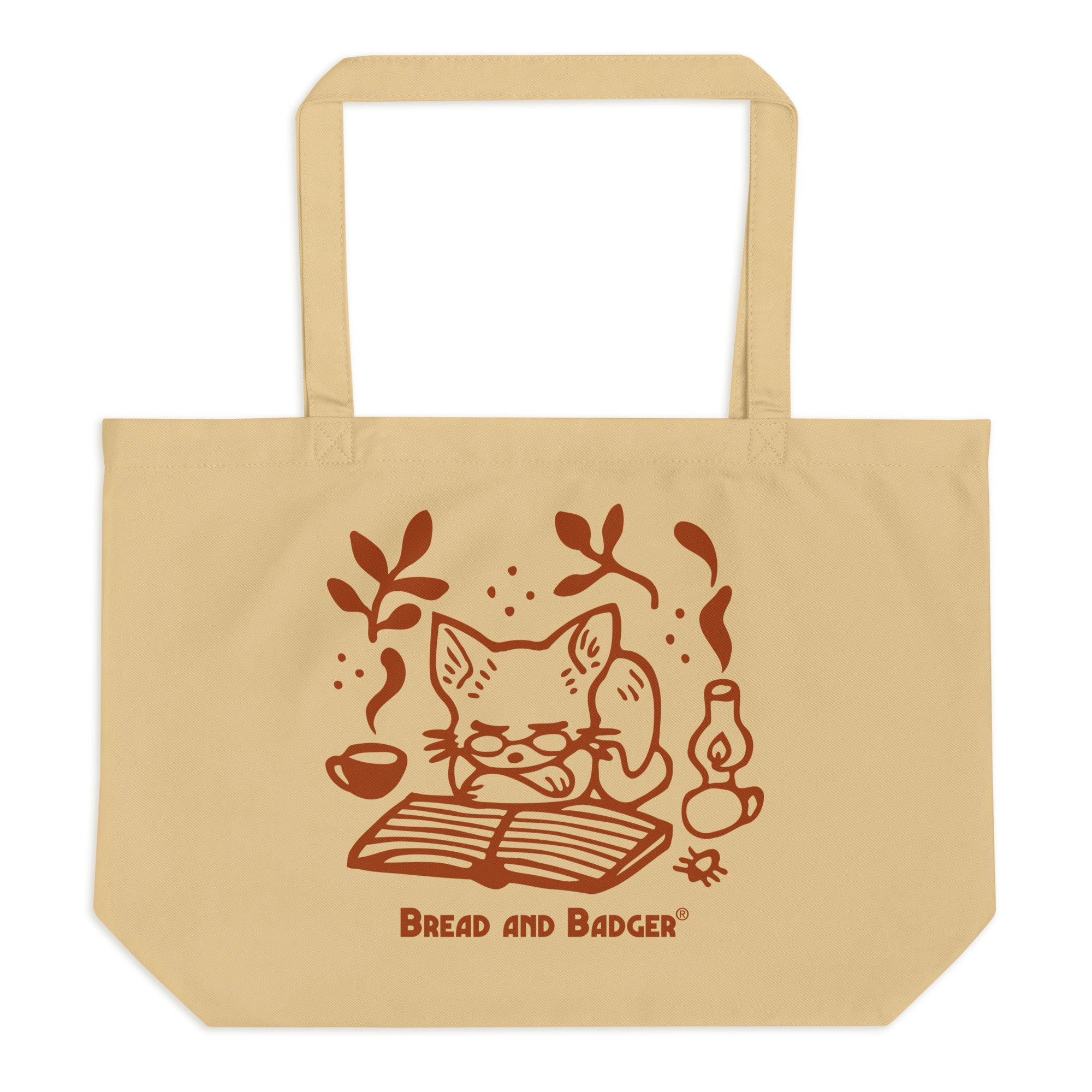 Beige tote bag with a red cat illustration and 'Bread and Badger' branding on a white background. The cat is sleeping in front of an open book, next to a steaming mug and lantern.