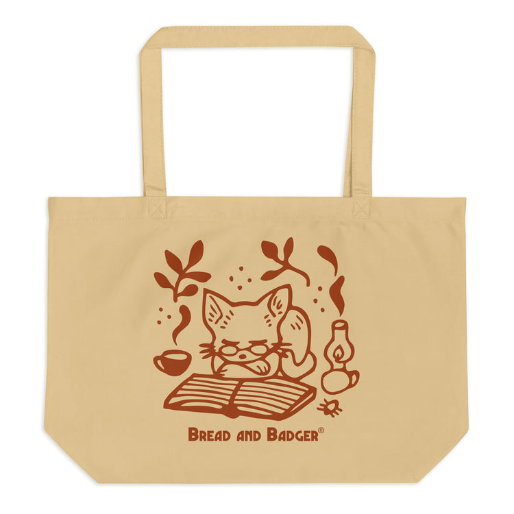 Beige tote bag with a red cat illustration and 'Bread and Badger' branding on a white background. The cat is sleeping in front of an open book, next to a steaming mug and lantern.
