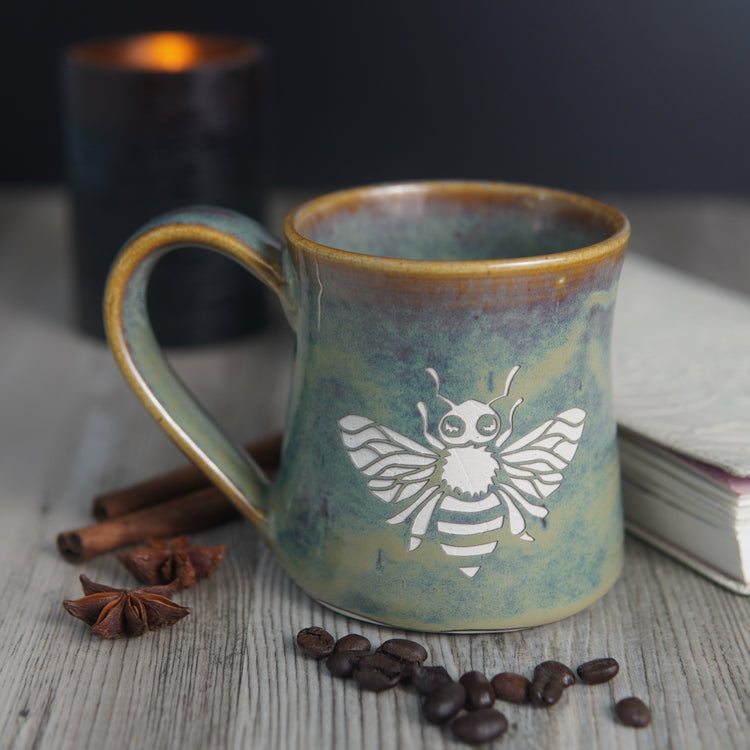 pottery mug engraved with a honey bee, next to a lit candle, book, coffee beans, cinnamon sticks, and star anise