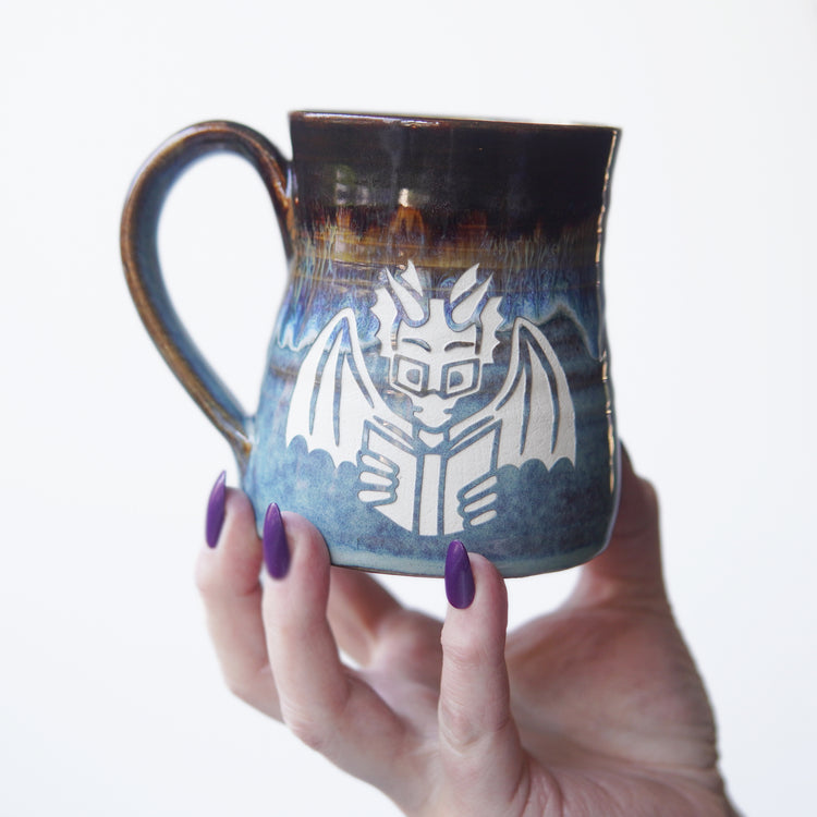 blue and brown drippy mug engraved with a winged dragon wearing glasses and reading a book