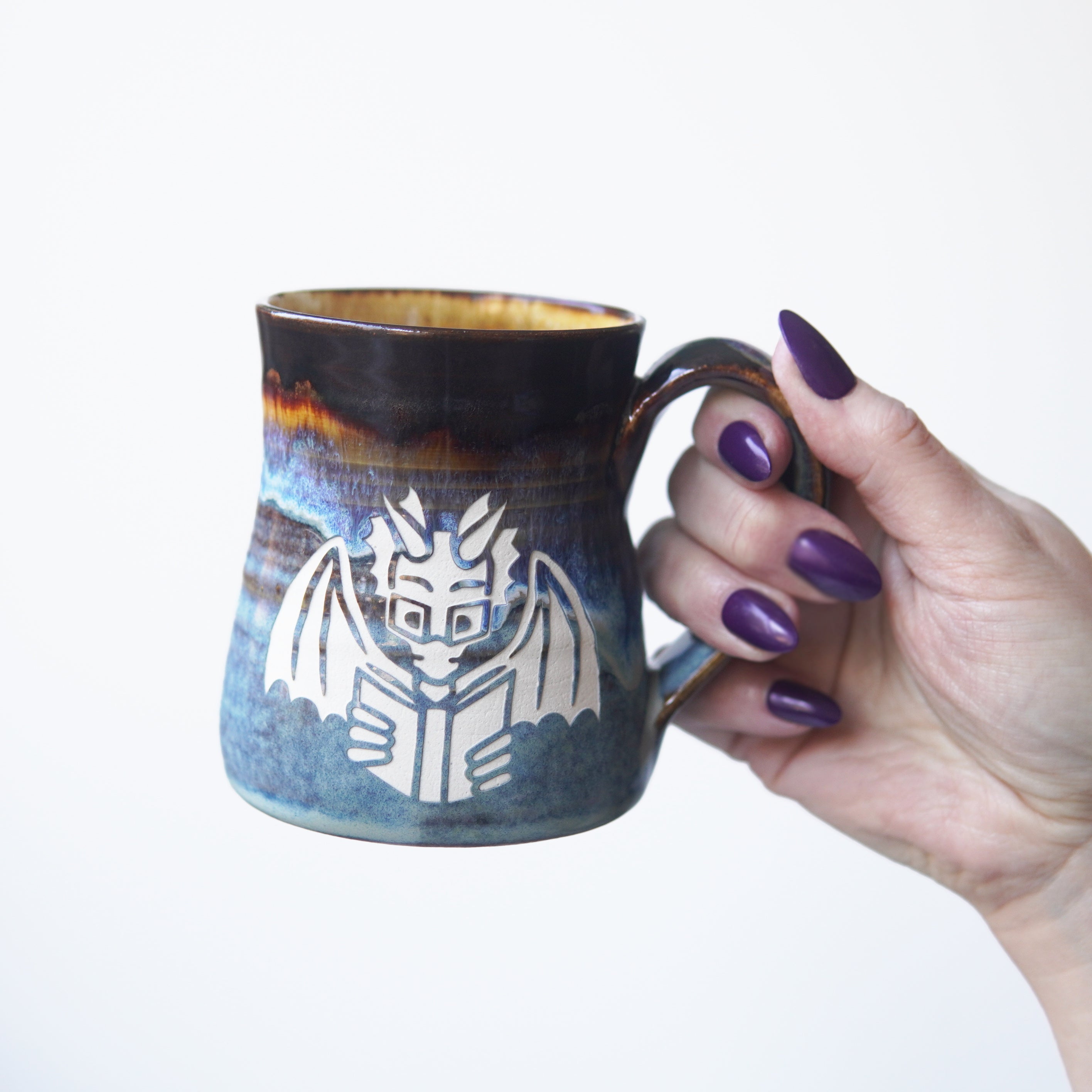 blue and brown drippy mug engraved with a winged dragon wearing glasses and reading a book