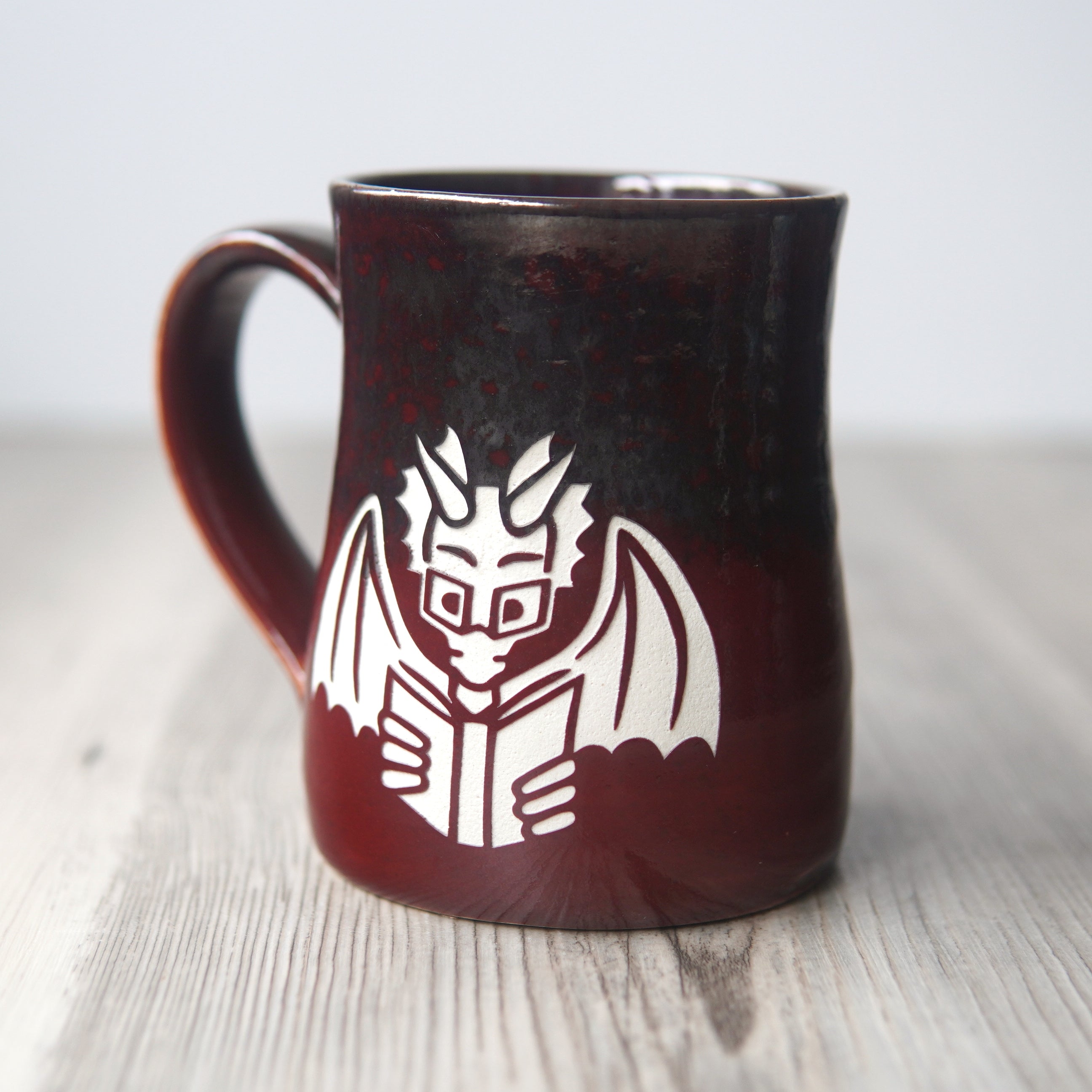 red and gray mug engraved with a winged dragon wearing glasses and reading a book