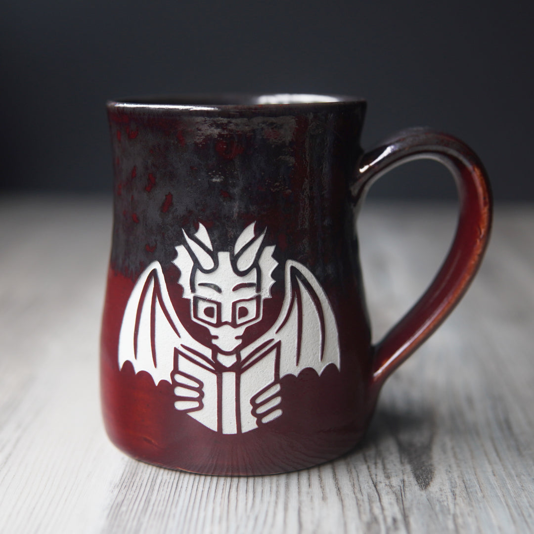 red and gray mug engraved with a winged dragon wearing glasses and reading a book