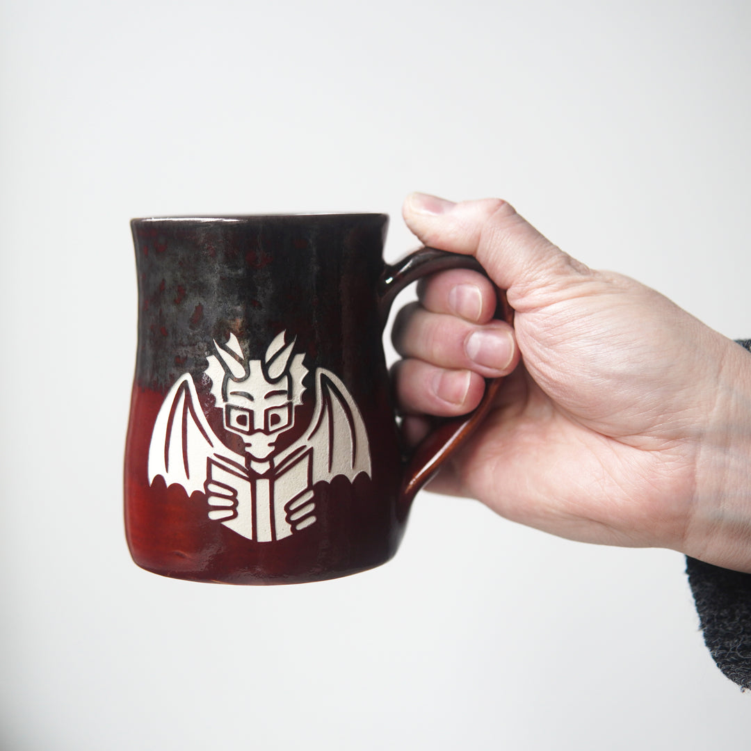 red and gray mug engraved with a winged dragon wearing glasses and reading a book