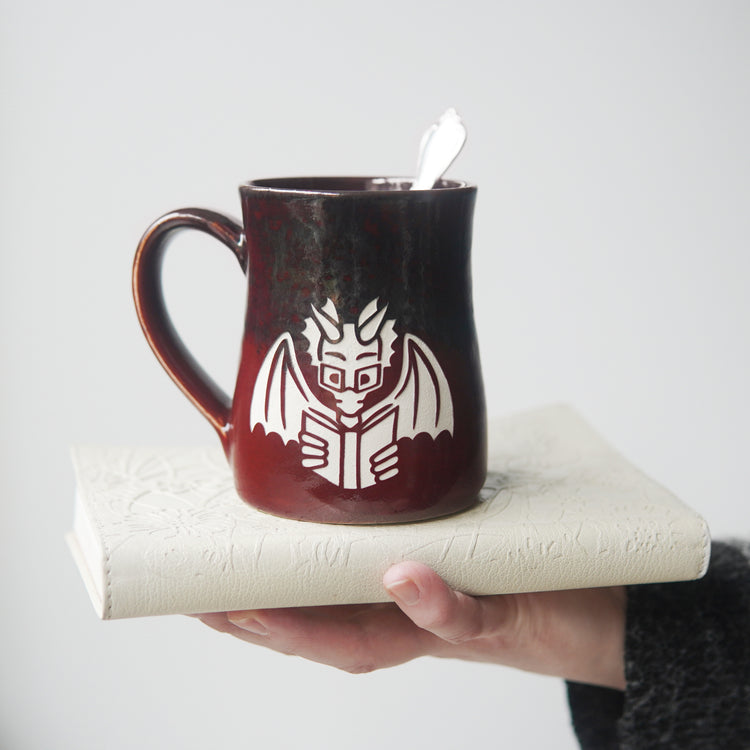 red and gray mug engraved with a winged dragon wearing glasses and reading a book