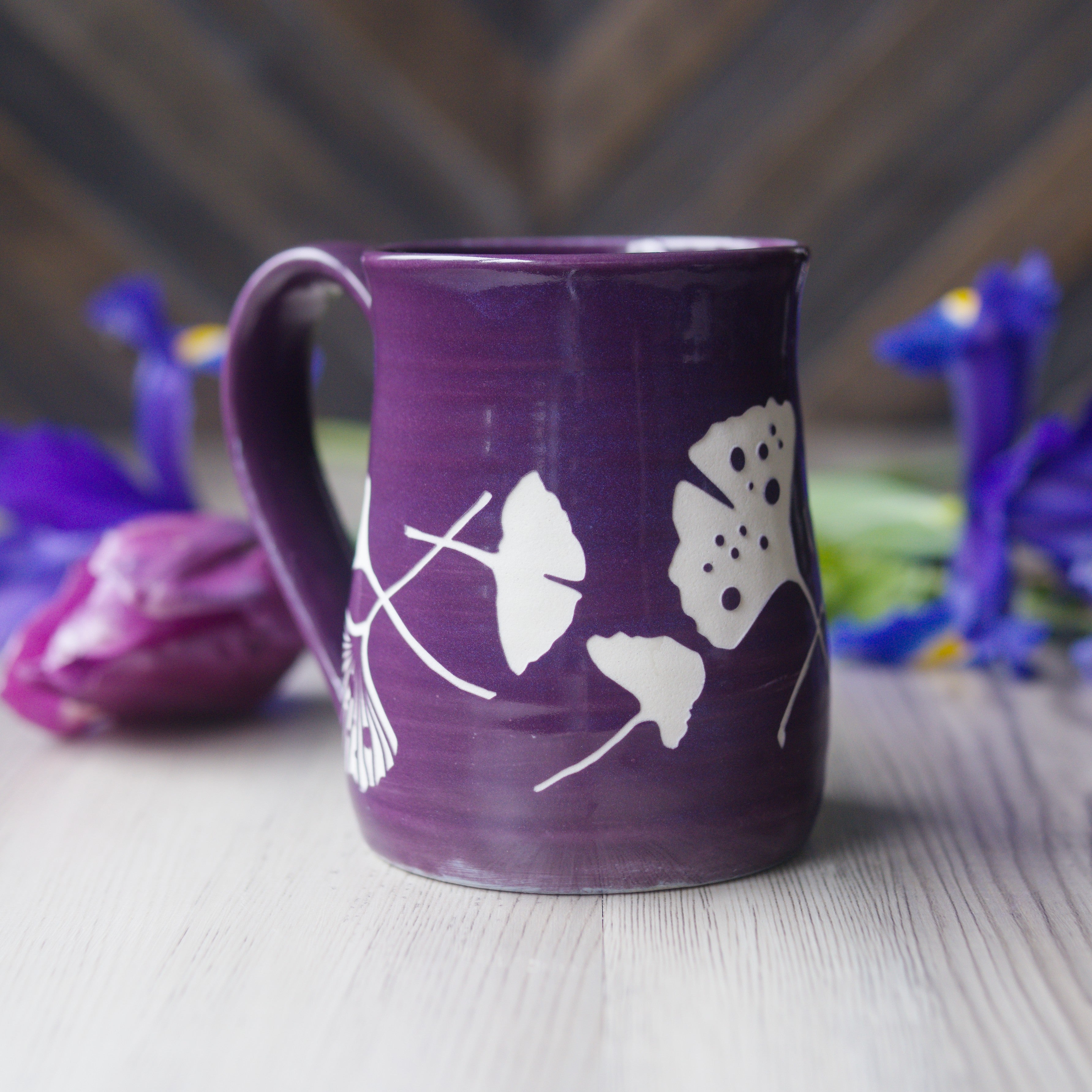 Purple mug with white floral designs on a wooden surface with flowers in the background