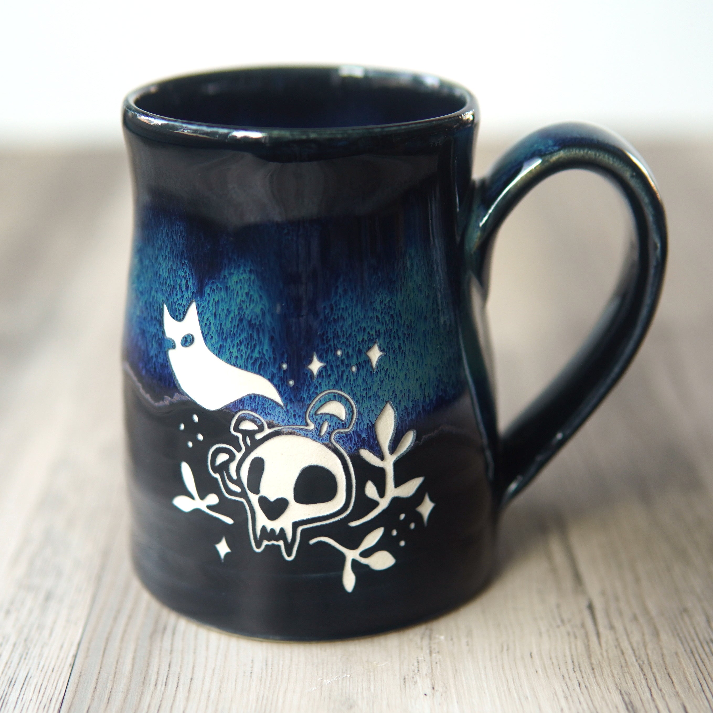 A dark blue and black glazed ceramic mug with a handle, decorated with a white skull, cat ghost, and stars design, resting on a light wood surface.