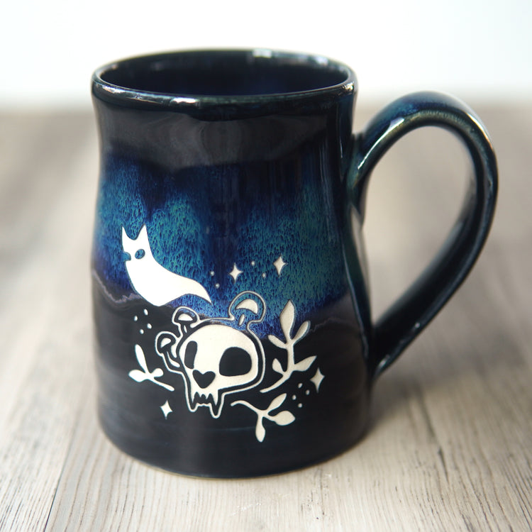A dark blue and black glazed ceramic mug with a handle, decorated with a white skull, cat ghost, and stars design, resting on a light wood surface.